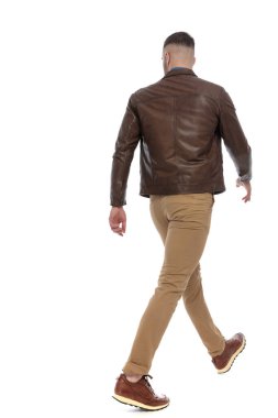 back view picture of cool casual man in brown leather jacket, chino pants and sneakers looking away and walking in front of white background in studio