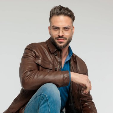 Portrait of handsome casual man posing with a subtle smile, squatting, wearing eyeglasses against gray studio background
