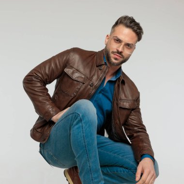 Portrait of young casual man resting his arms and posing sexy, squatting, wearing eyeglasses against gray studio background