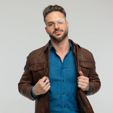 Portrait of handsome casual man arranging his leather jacket, standing, wearing eyeglasses against gray studio background