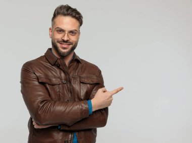 Portrait of young casual man pointing to the side and smiling, standing, wearing eyeglasses against gray studio background