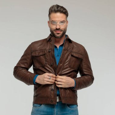 Portrait of young casual man closing his jacket with style, standing, wearing eyeglasses against gray studio background