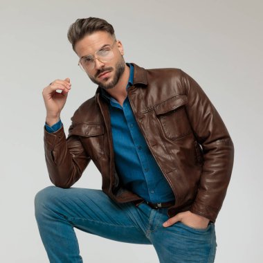 Portrait of handsome casual man putting hand in pocket and posing with attitude, leaning on one leg, wearing eyeglasses against gray studio background