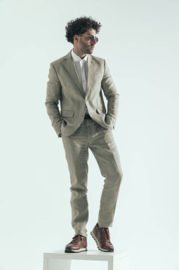 sexy brunette businessman with curly hair looking to side while posing with hands in pockets on grey background in studio