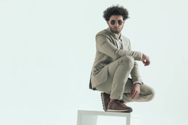 full body picture of iranian man with curly hair holding elbow on knee while crouching on white chair in front of grey background in studio
