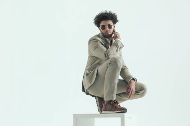 turkish young man with curly hair wearing sunglasses crouching and holding elbow on knee while posing in a fashion way on greybackground