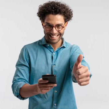 Portrait of attractive casual man texting on his phone and giving a like, standing, wearing a shirt and eyeglasses against gray studio background