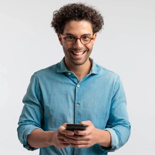 携帯電話で楽しいテキストメッセージを持っているハンサムなカジュアルな男の肖像画 立って 灰色のスタジオの背景にシャツと眼鏡をかけて — ストック写真