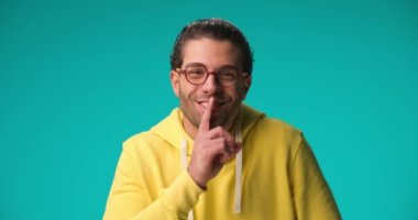 happy casual man with glasses in yellow hoodie laughing, holding finger to mouth and making silence on blue background