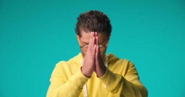 young Arabic guy with glasses holding hands together, praying, pointing fingers to the sky on blue background