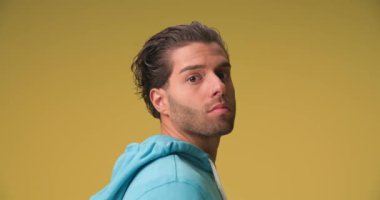 side view of sexy man with curly hair in blue hoodie looking to side, holding finger to mouth and making silence gesture on yellow background