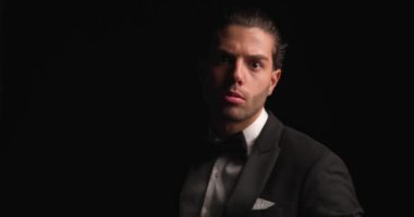 project video of sexy Lebanese man adjusting elegant black tuxedo and bowtie, making a hand gesture and inviting before walking away in darkness
