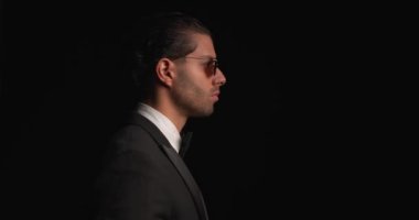 side view of attractive young businessman wearing black tuxedo and sunglasses standing and looking to side in front of black background