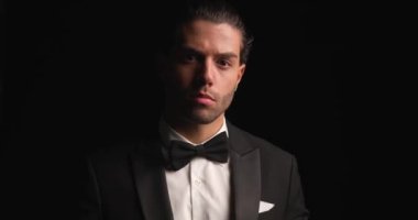 sexy Arabic man fixing tuxedo and bowtie, smiling while pointing finger and inviting in front of black background