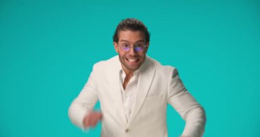 enthusiastic man with glasses wearing white suit being surprised, celebrating victory, dancing and enjoying the success on blue background