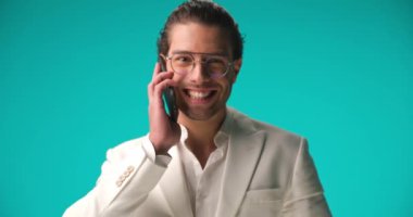 excited young man wearing white suit with open collar shirt, having a phone conversation, smiling, laughing and pointing fingers on blue background in studio