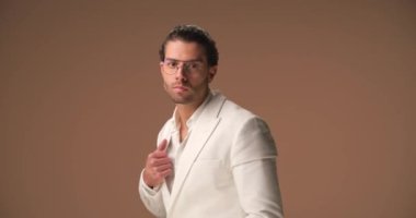 confident man in white suit with sunglasses and open collar shirt moving and posing in front of brown background in studio