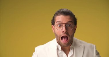 shocked Greek man with curly hair and eyeglasses opening mouth, screaming and being surprised and excited about the news in front of yellow mustard background