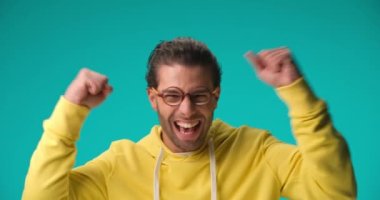 enthusiastic casual guy in yellow hoodie with glasses holding fists in the air and cheering the victory, pointing fingers and enjoying the success on blue background