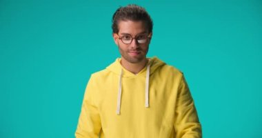excited casual guy with glasses wearing yellow hoodie, being happy, laughing and pointing fingers on blue background in studio