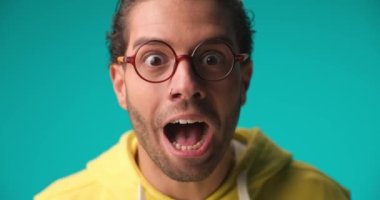 close up video of surprised young man in yellow hoodie with glasses celebrating the victory and enjoying the success on blue background