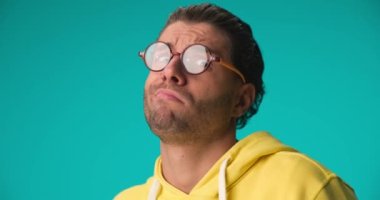 silly young man with curly hair and glasses being foolish, making faces and having fun, sticking out tongue and laughing on blue background