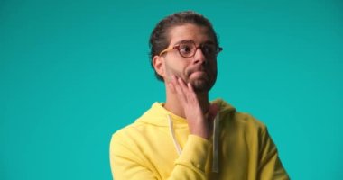casual Greek man in yellow hoodie with glasses looking around, holding his head and being bored and tired on blue background