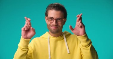 happy curly hair guy in yellow hoodie crossing fingers for good luck, wishing and hoping for the best on blue background