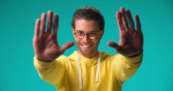 青い背景の前で腕を上げて笑顔で額を組んでいる黄色のスウェットシャツの幸せなトルコ人のビデオ — ストック動画