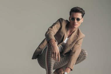 happy man with sunglasses wearing beige jacket and plaid pants and crouching in front of grey background in studio