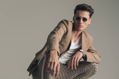 fashion man with plaid pants and beige jacket posing while crouching with elbows on knees in front of grey background in studio