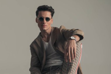 happy confident man wearing sunglasses and beige jacket with plaid pants holding elbow on knee and posing while sitting on grey background
