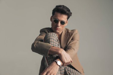 cool guy wearing plaid pants with beige jacket and sunglasses posing with knee up in front of grey background in studio