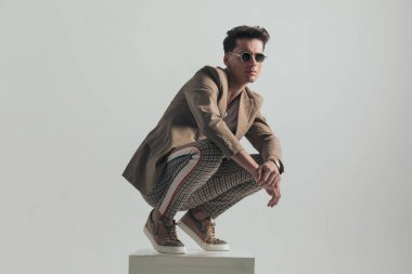 cool fashion model with plaid pants and beige jacket crouching on top of white box and posing with elbows above knees in front of grey background in studio