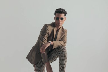 confident man with sunglasses holding knee up with elbow above and posing in a cool way in front of grey background in studio