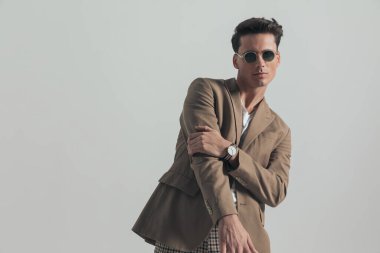 portrait of cool casual man with beige jacket and sunglasses holding arms in fashion pose and posing in front of grey background in studio