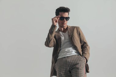 sexy young man looking away, fixing glasses and posing with hand in pocket in front of grey background in studio