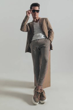 cool fashion man fixing glasses while laying on box with hand in pocket and looking away in front of grey background in studio, full body