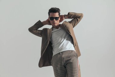attractive young man with sunglasses holding hands behind neck and posing in a cool way on grey background in studio