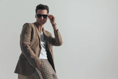 side view of cool fashion guy with hand in pocket adjusting sunglasses while laying on a box in front of grey background in studio