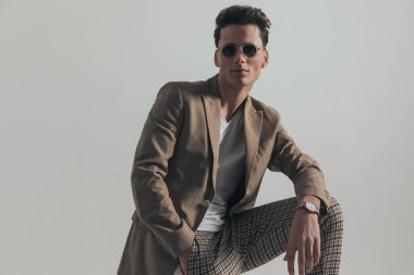 handsome guy with dark hair holding knee up with elbow above posing with hand in pocket in front of grey background in studio