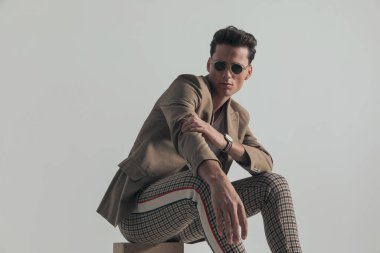 casual young man with plaid pants adjusting jacket sleeve and posing while sitting in front of grey background in studio