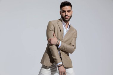 Portrait of attractive businessman holding his elbow and posing with attitude, standing, against gray studio background