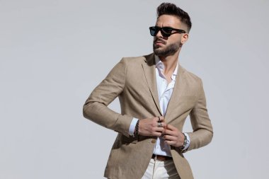 Portrait of sexy businessman looking away and closing his suit in fashion pose, standing, wearing sunglasses against gray studio background