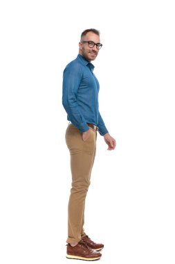 side view of casual nerdy guy in denim shirt standing in line with hand in pocket in front of white background in studio