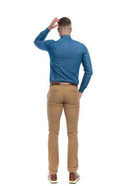 full body picture of confused young guy in denim shirt scratching head and thinking in front of white background in studio