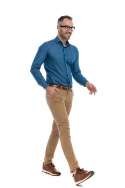 side view of handsome young man in denim shirt smiling and walking while looking to side and holding hand in pocket on white background