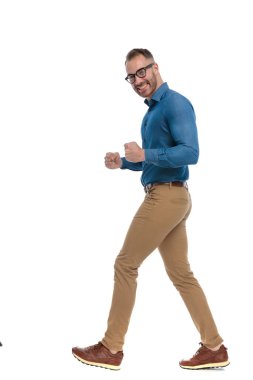 side view of excited guy with glasses celebrating victory and walking in front of white background in studio