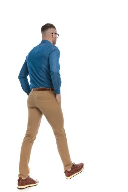 rear view of handsome casual guy in denim shirt walking with  hands in pockets on white background in studio