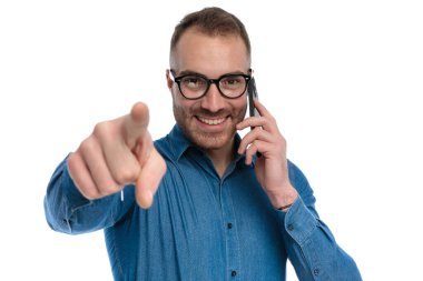 attractive bearded guy with glasses talking on the phone and pointing finger, smiling and posing on white background in studio